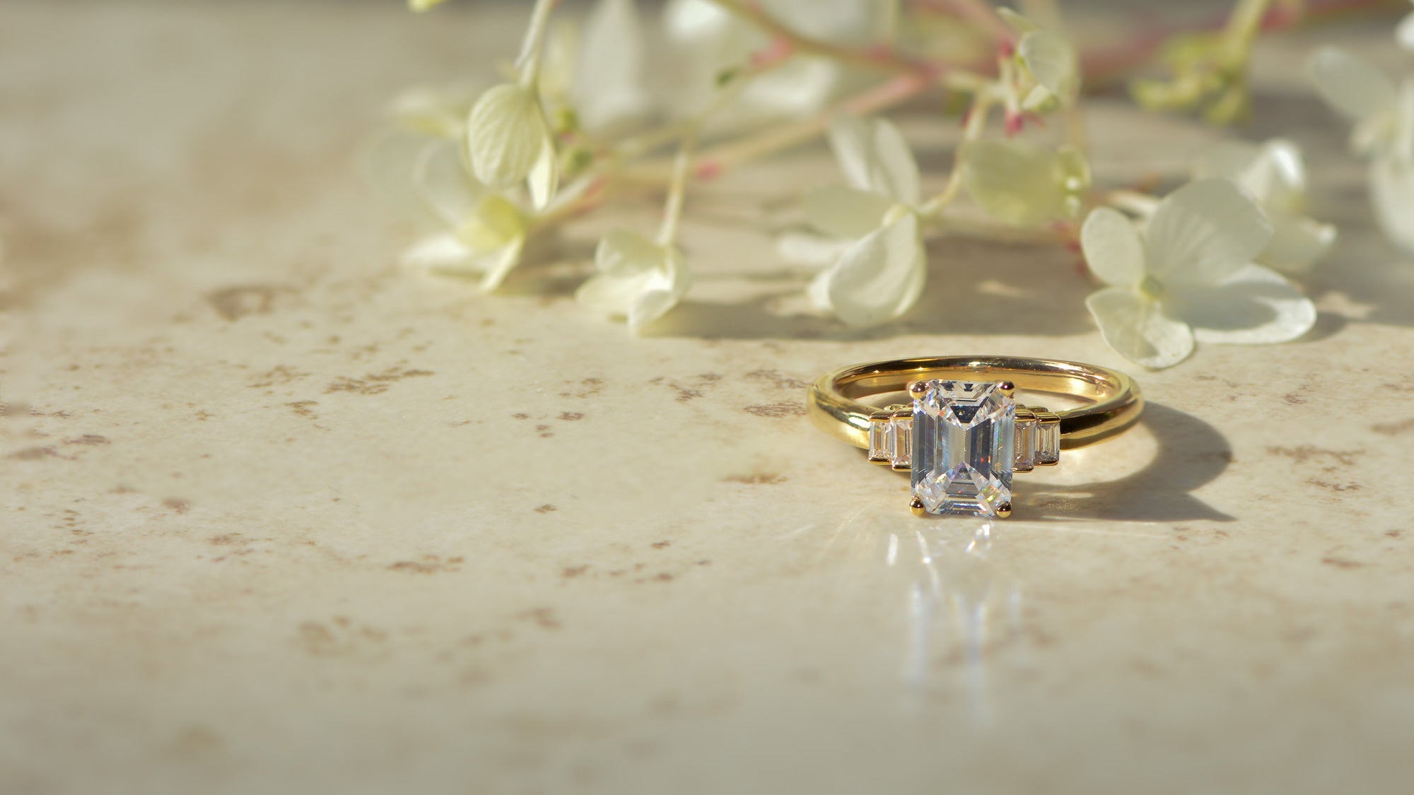 Emerald cut art deco inspired diamond engagement ring on a marble surface with blurred flowers in the background