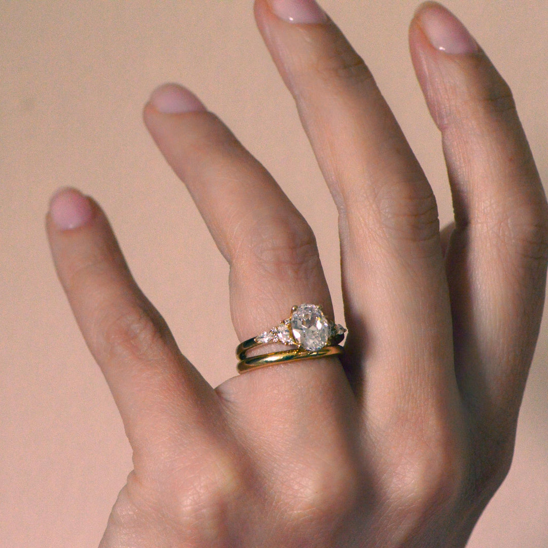 Hand wearing a gold and diamond ring with an oval cut centre stone on a beige background