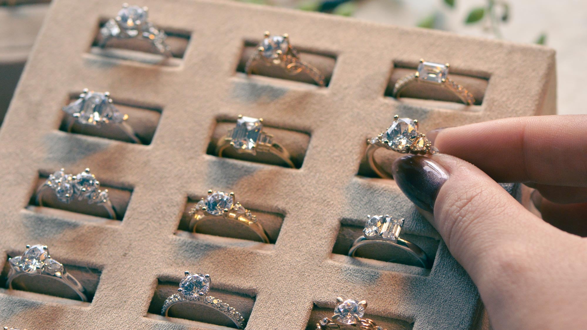 Hand holding a diamond ring above a display of similar rings in a box.