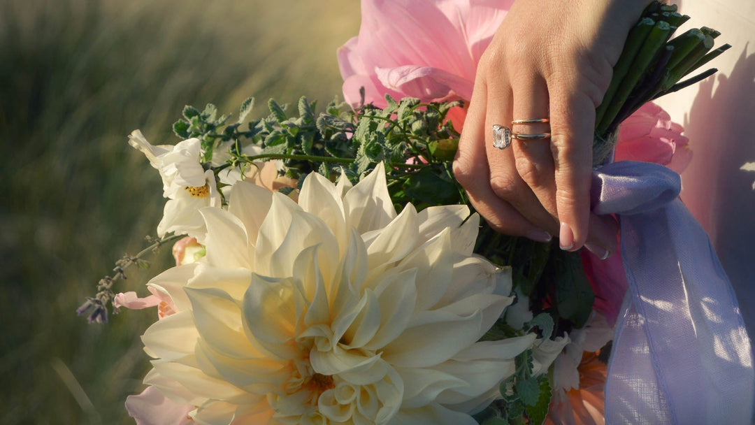 Close up of a hand holding a bouquet of flowers in neutral colours. There is a lab diamond engagement ring and wedding band worn on the hand.