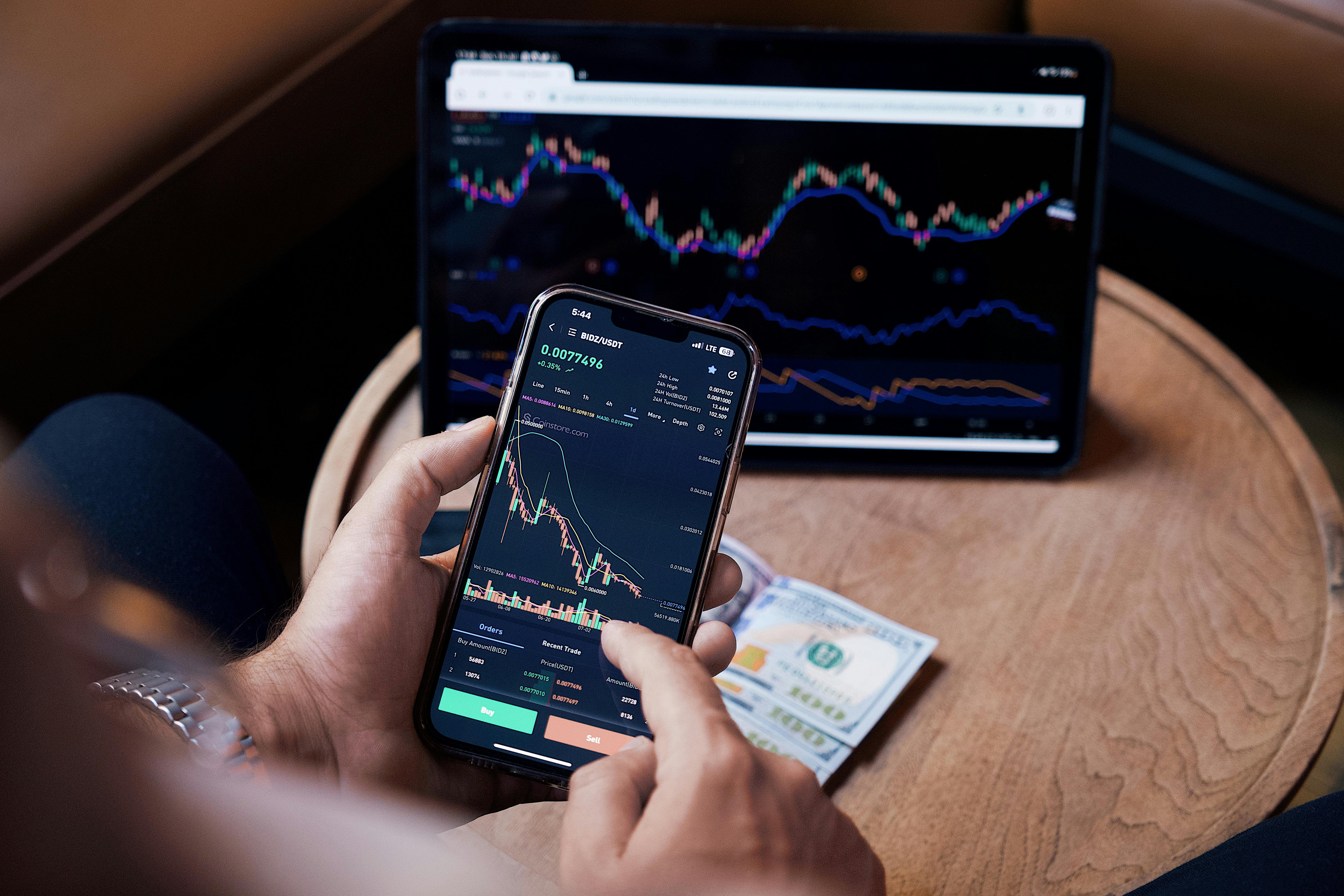 A person holding a mobile phone with a computer tablet in the background. Both screen show financial graphs of stock and shares 