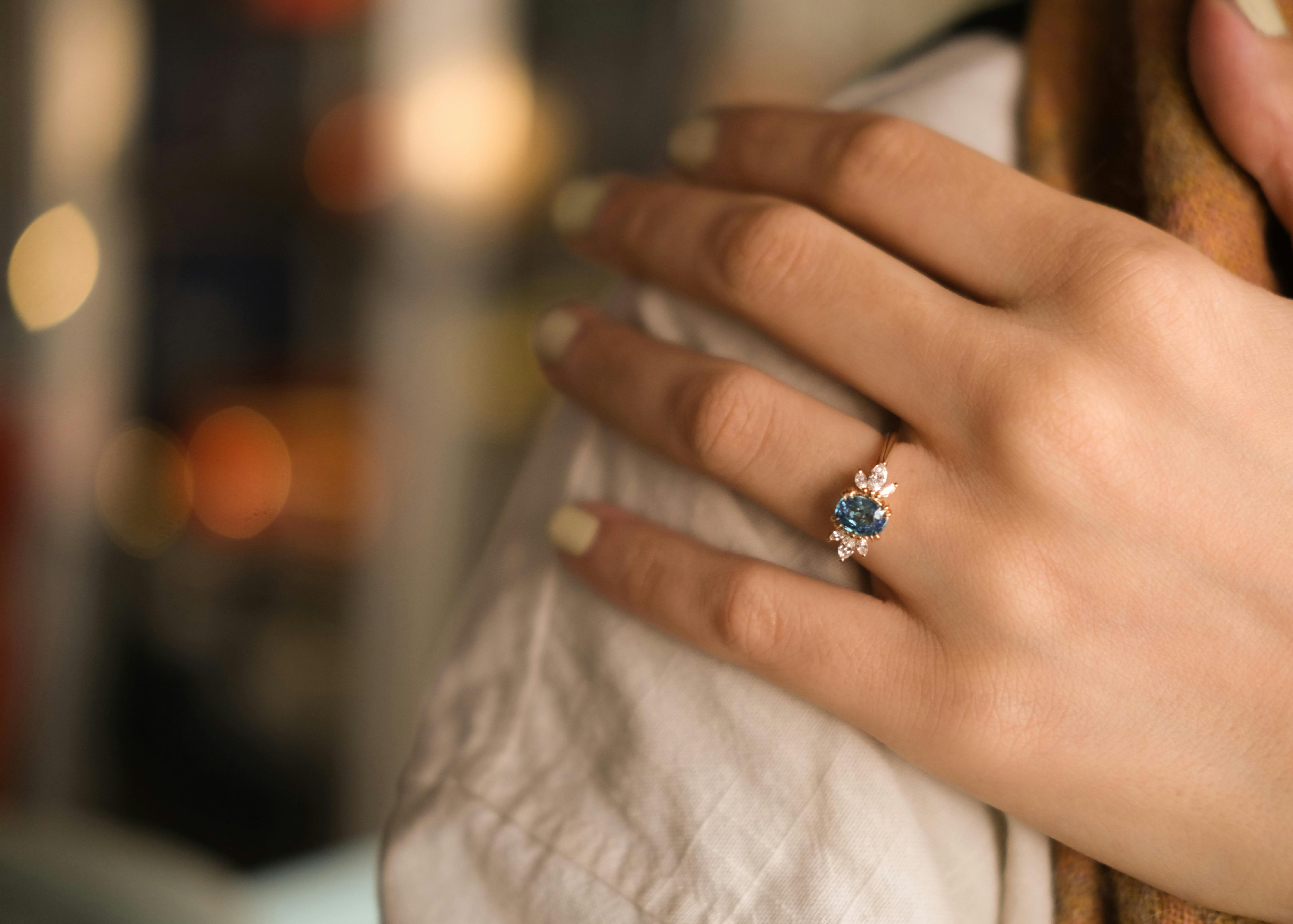 A gold engagement ring with a bright blue centre stone and marquise cut side stones on a woman's hand. She rests her hand on her shoulder
