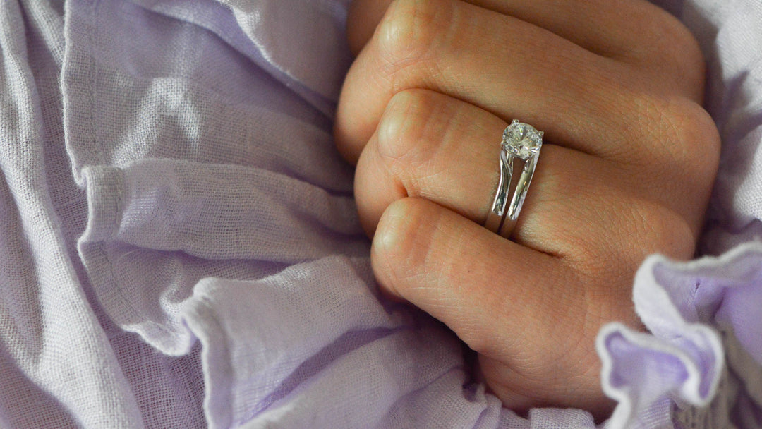 Round brilliant cut solitaire diamond engagement ring worn on the hand worn with a platinum wedding band with lilac linen in the background