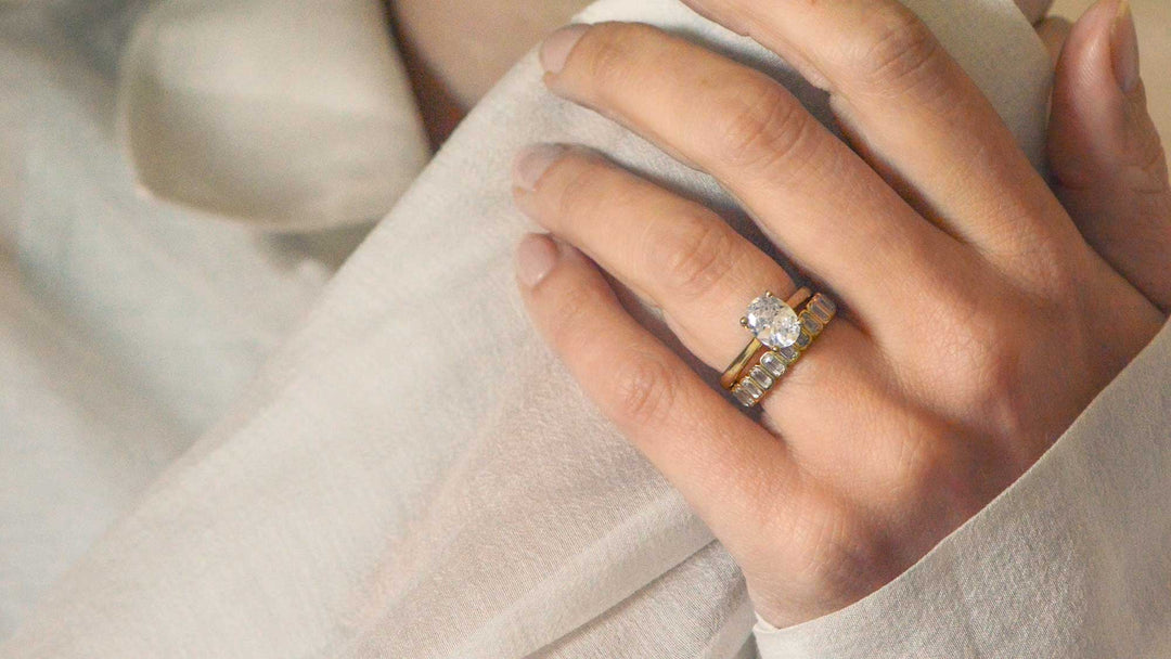 Oval cut solitaire engagement ring worn on the hand with an gold wedding band, bezel set with emerald cut diamonds on a pale background