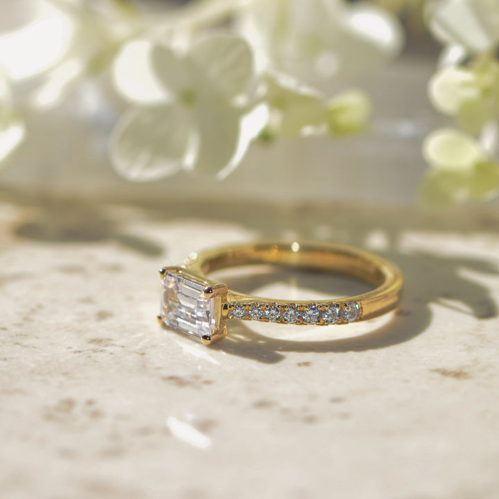 Gold ring with an emerald cut diamond on a marble surface with flowers in the background