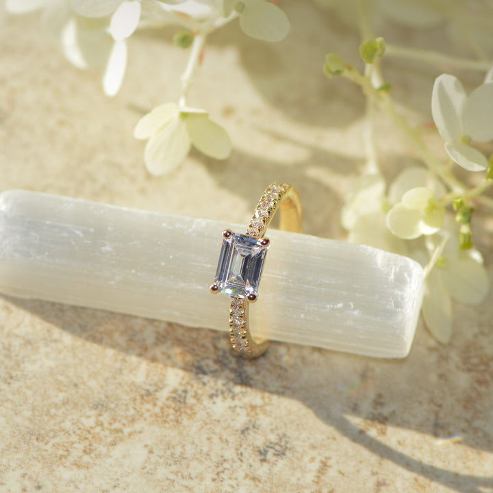 Gold engagement ring with an emerald cut diamond on a white stone with flowers in the background