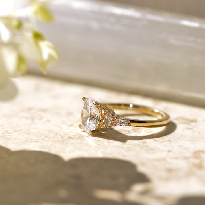 Gold ring with an oval diamond on a textured surface with flowers in the background