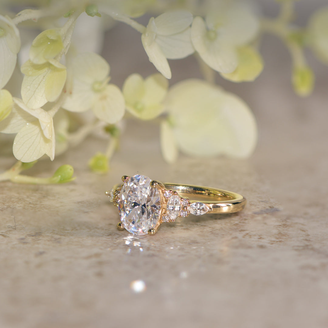 Gold oval cut diamond engagement ring on a textured surface with blurred flowers in the background
