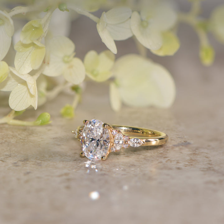 Gold oval cut diamond engagement ring on a textured surface with blurred flowers in the background