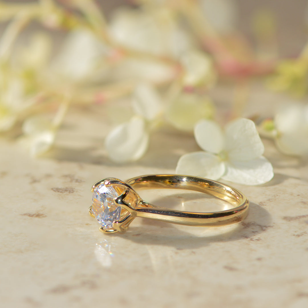 Gold, round brilliant cut diamond engagement ring on a marble surface with flowers in the background