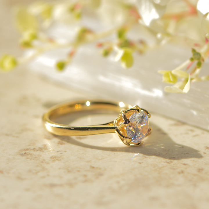 Gold six claw engagement ring with a round cut diamond set in a floral inspired tulip setting on a marble surface with flowers in the background