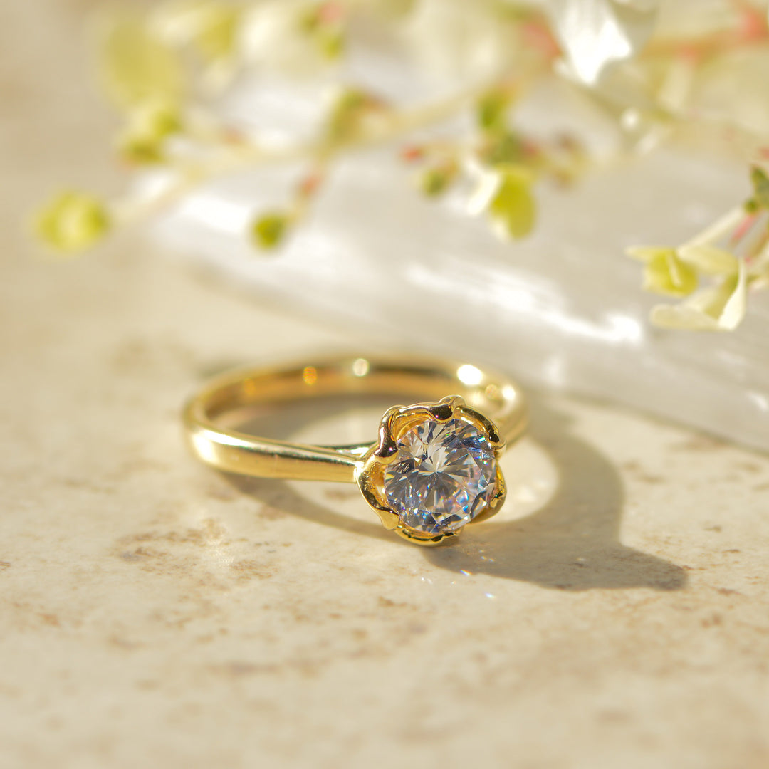 Gold solitaire ring with a round diamond on a light surface with flowers in the background