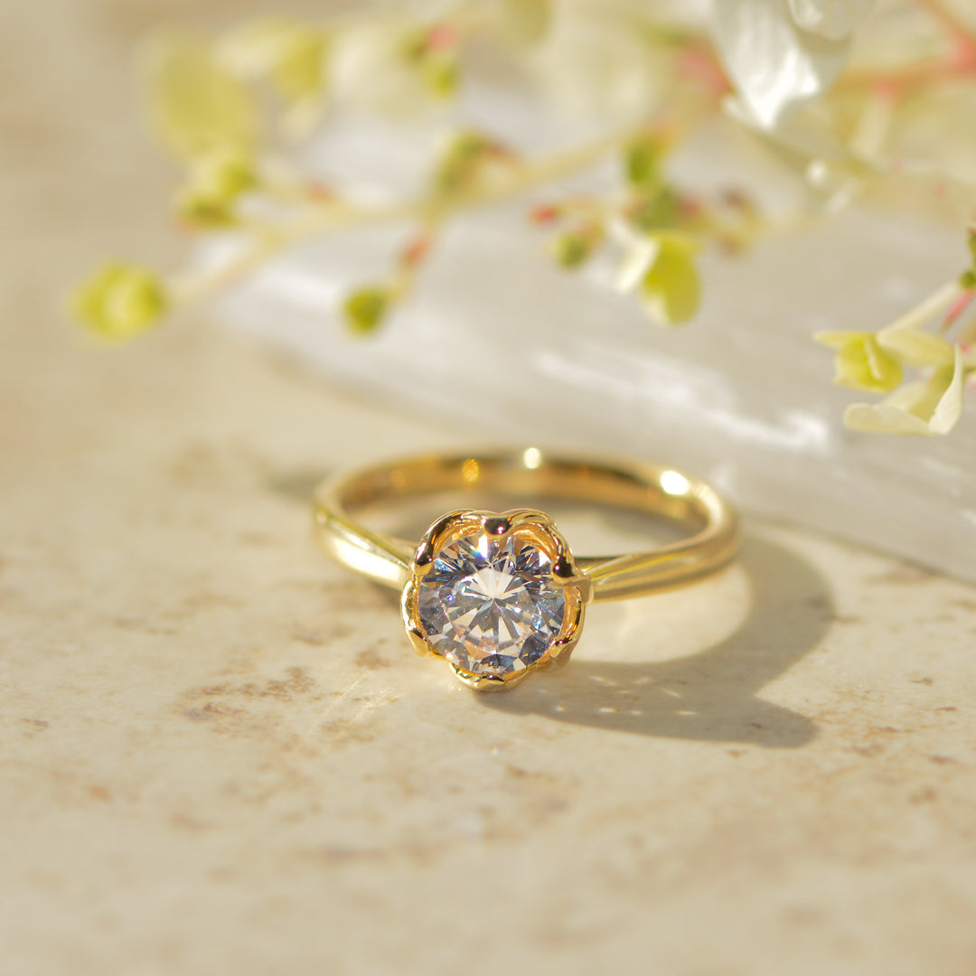 Gold solitaire engagement ring with a round brilliant cut diamond on a marble surface with floral background