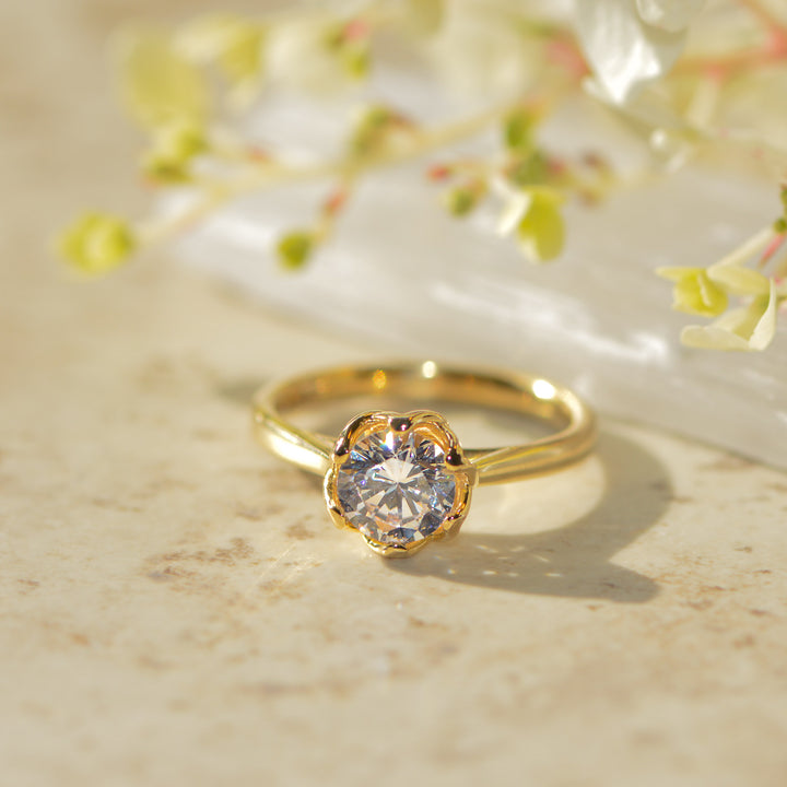 Gold solitaire engagement ring with a round brilliant cut diamond on a marble surface with floral background