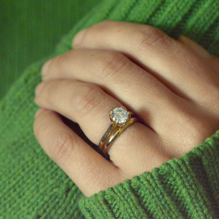 Gold solitaire engagement ring with a round brilliant cut diamond on a hand against a green background