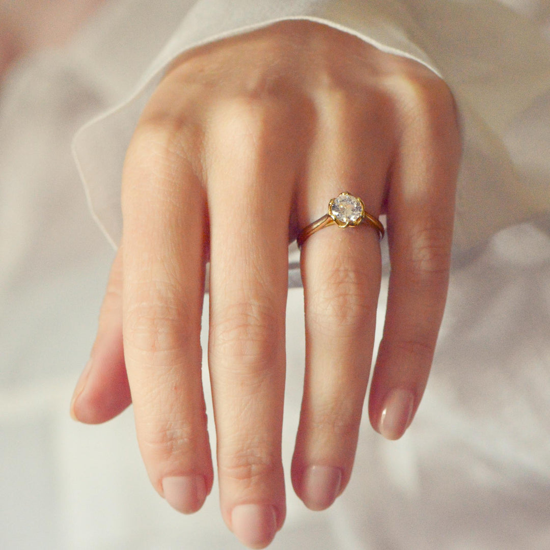 Gold solitaire engagement ring with a round brilliant cut diamond on a person's finger against a neutral background