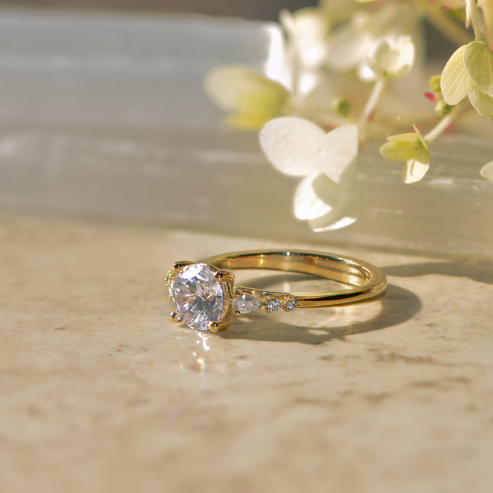 Gold engagement ring with diamond set shoulders on a marble surface with flowers in the background