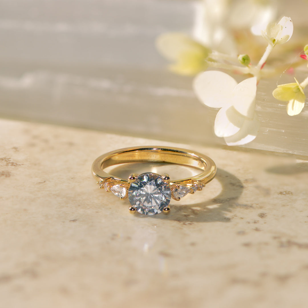 Gold round brilliant cut diamond ring on a marble surface with flowers in the background