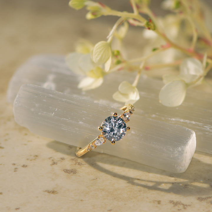 Gold round cut diamond ring with pear cut side stones on a white stone with greenery in the background