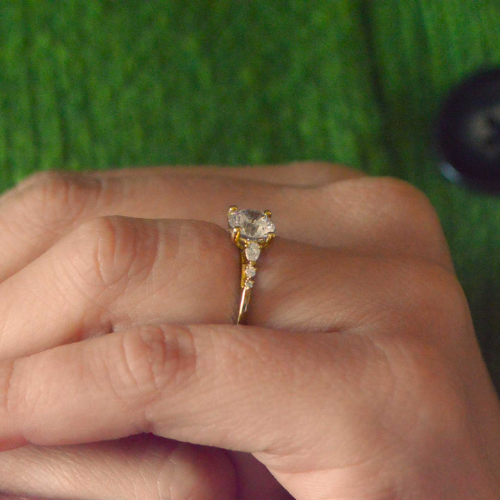 Gold engagement ring with a round brilliant cut diamond on a person's finger against a green background