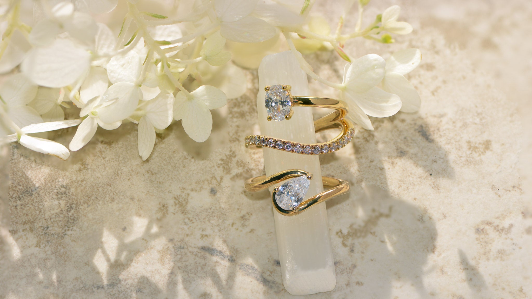 Two gold & diamond engagement rings on a white stone surface with flowers.