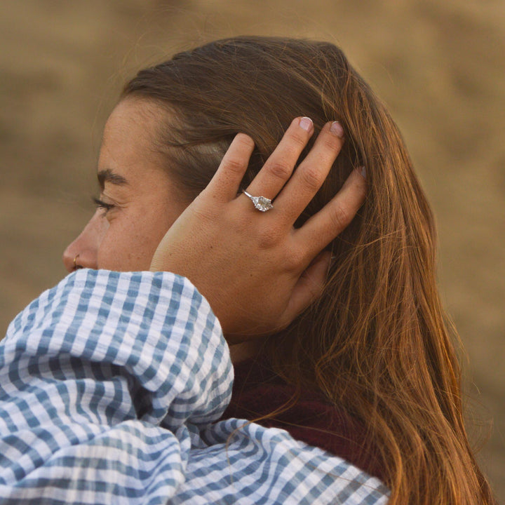 Woman with hand on head wearing a blue checkered shirt and Art Deco ring, against a blurred natural background