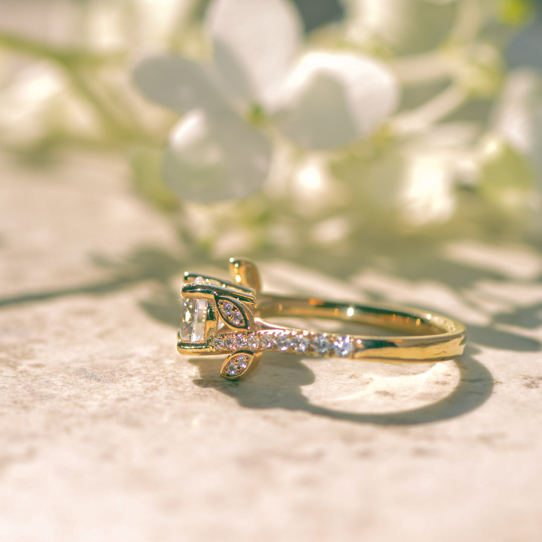 Gold shoulder set engagement ring with diamond accents on a light surface with blurred greenery