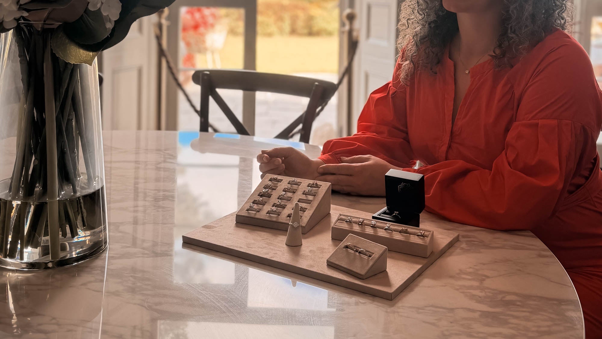A person in a bright red top sits at a marble table with a wooden jewelry tray holding small boxed items, alongside a black jewelry box and a large glass vase in a softly lit indoor setting.