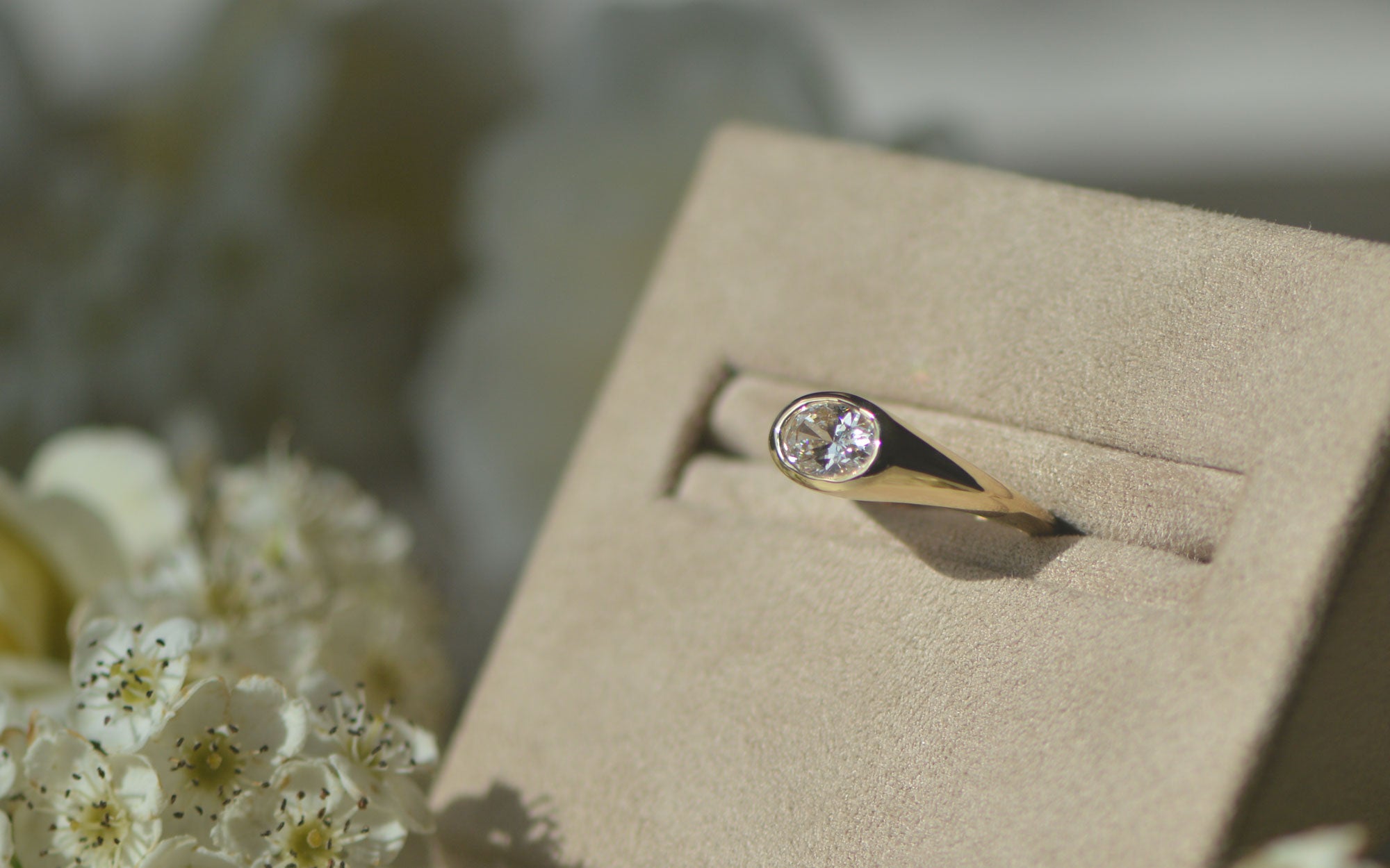 Bespoke gold engagement ring set with a oval cut diamond on a beige box with flowers in the background