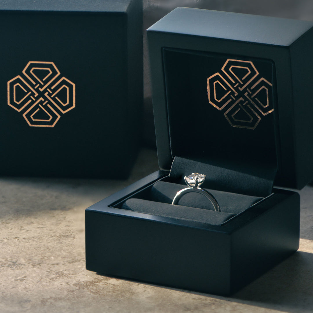 Diamond ring in an open black jewelry box with a brand logo on a textured surface