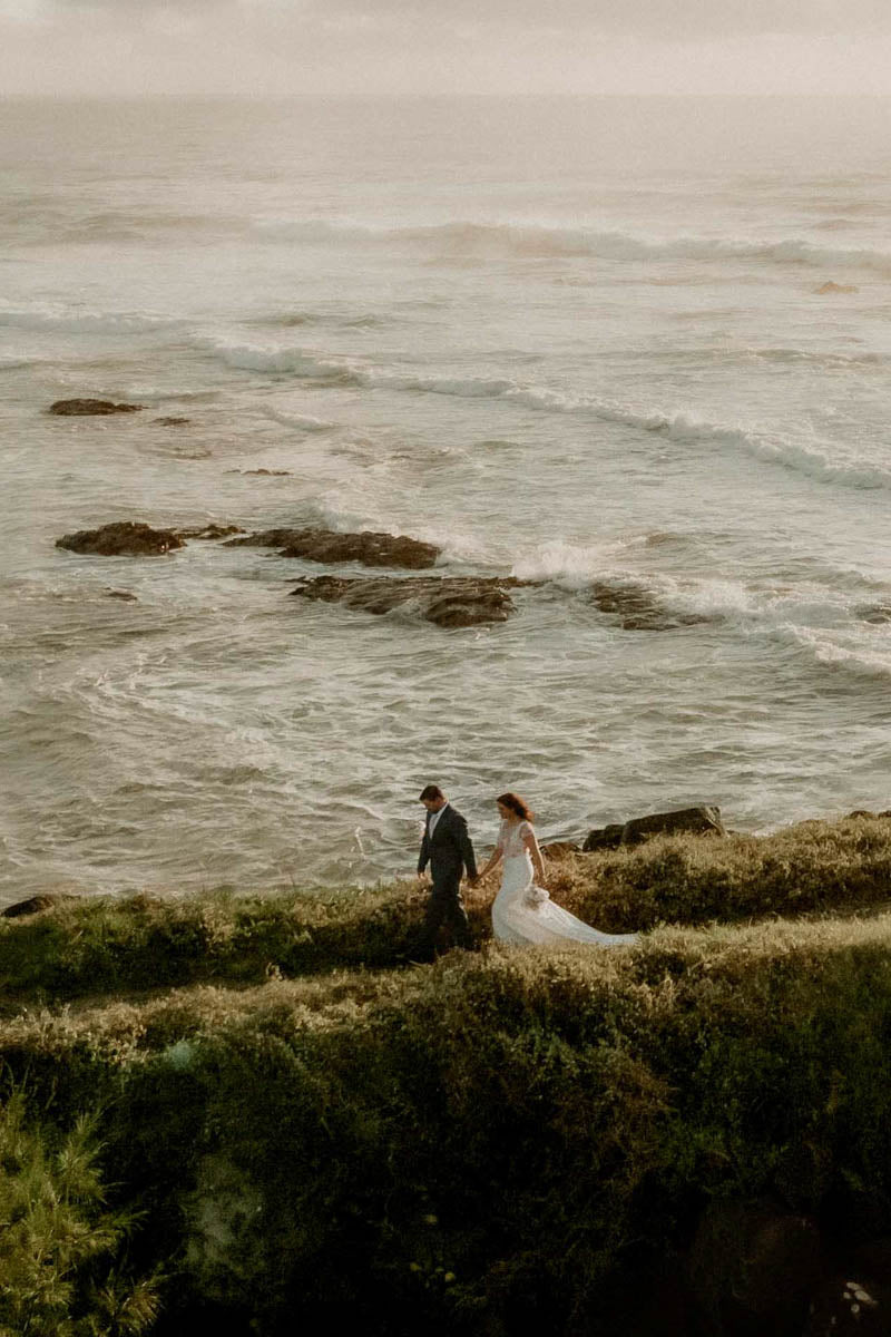 A bride and a groom walk hand in hand along a Cornish cliffside overlooking a rocky ocean coastline at golden hour.
