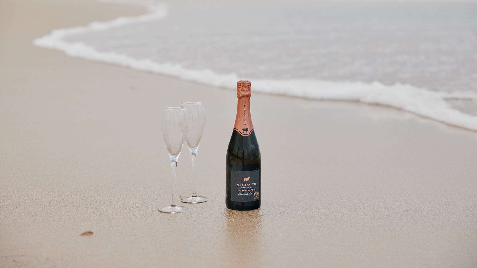 A bottle of Cornish champagne by the shoreline on a beach in Cornwall for a proposal celebration.