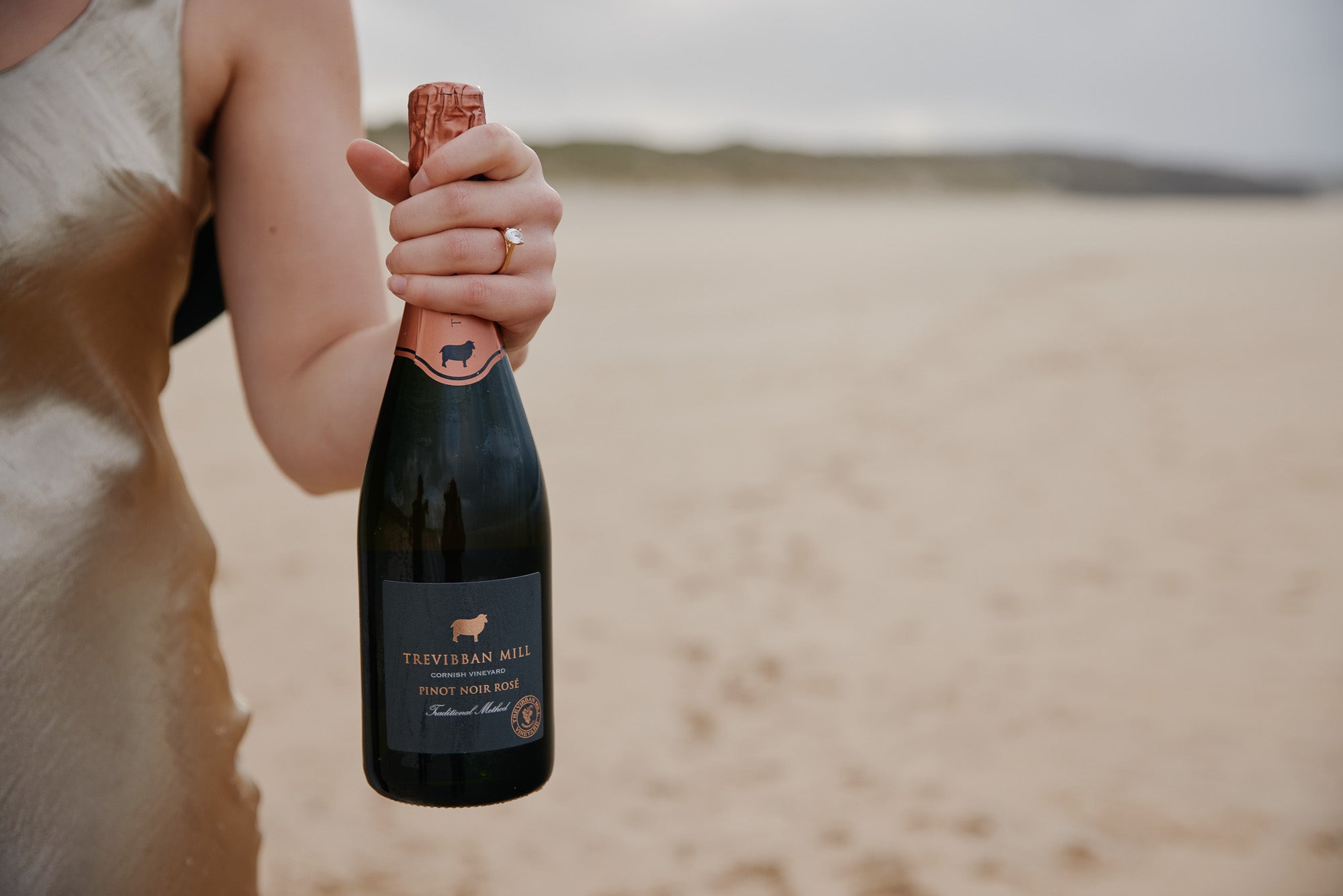 Person holding a bottle of Trevibban Mill wine on a beach with an engagement ring on their finger