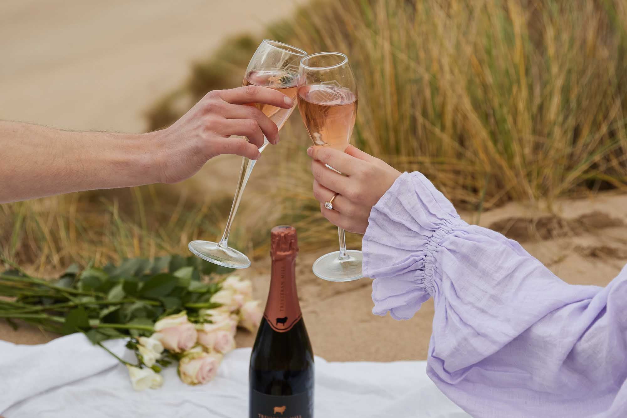 Two people clinking champagne glasses on a beach with a bottle of champagne and flowers.