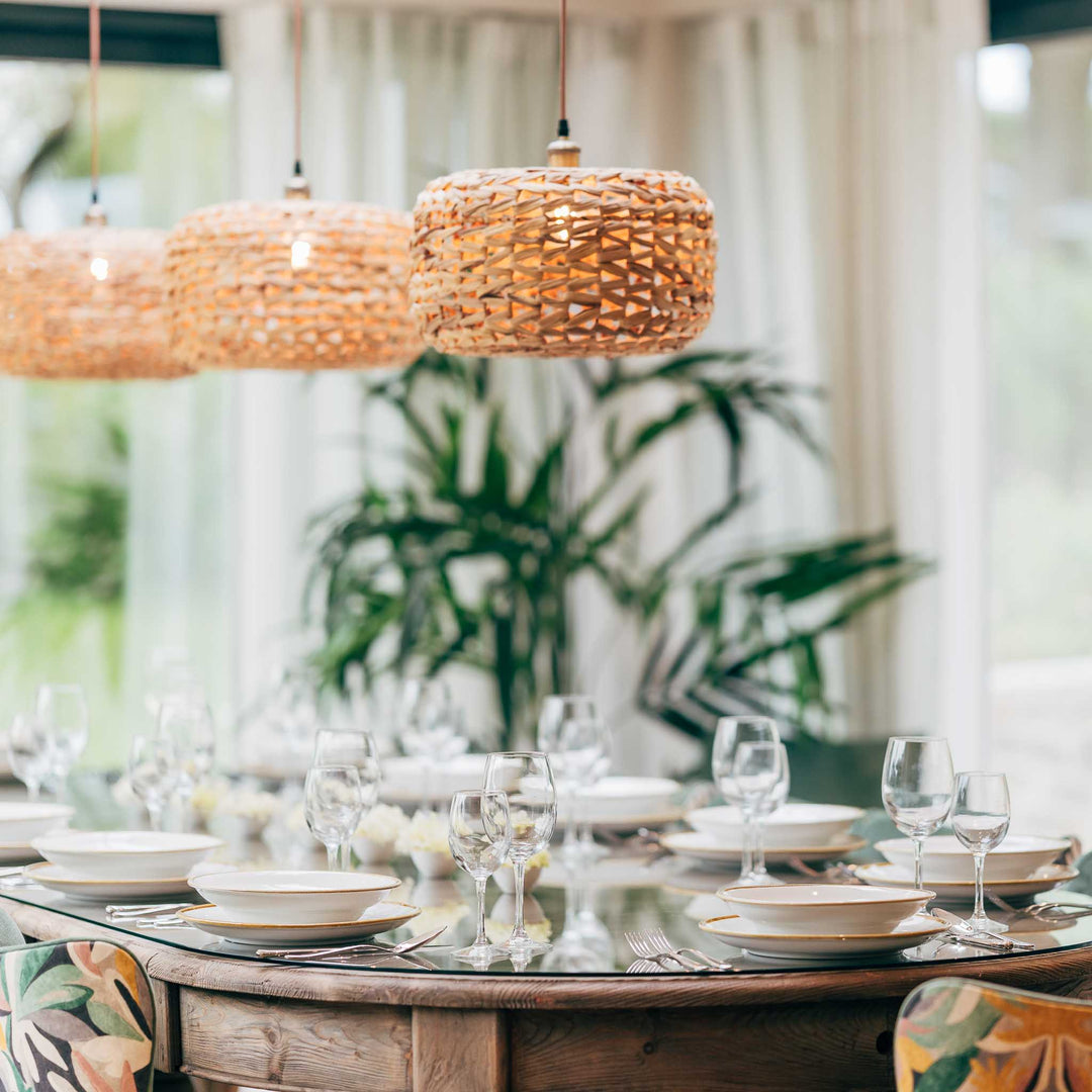 Dining table set with plates, glasses, and cutlery under wicker pendant lights with a blurred green plant background.