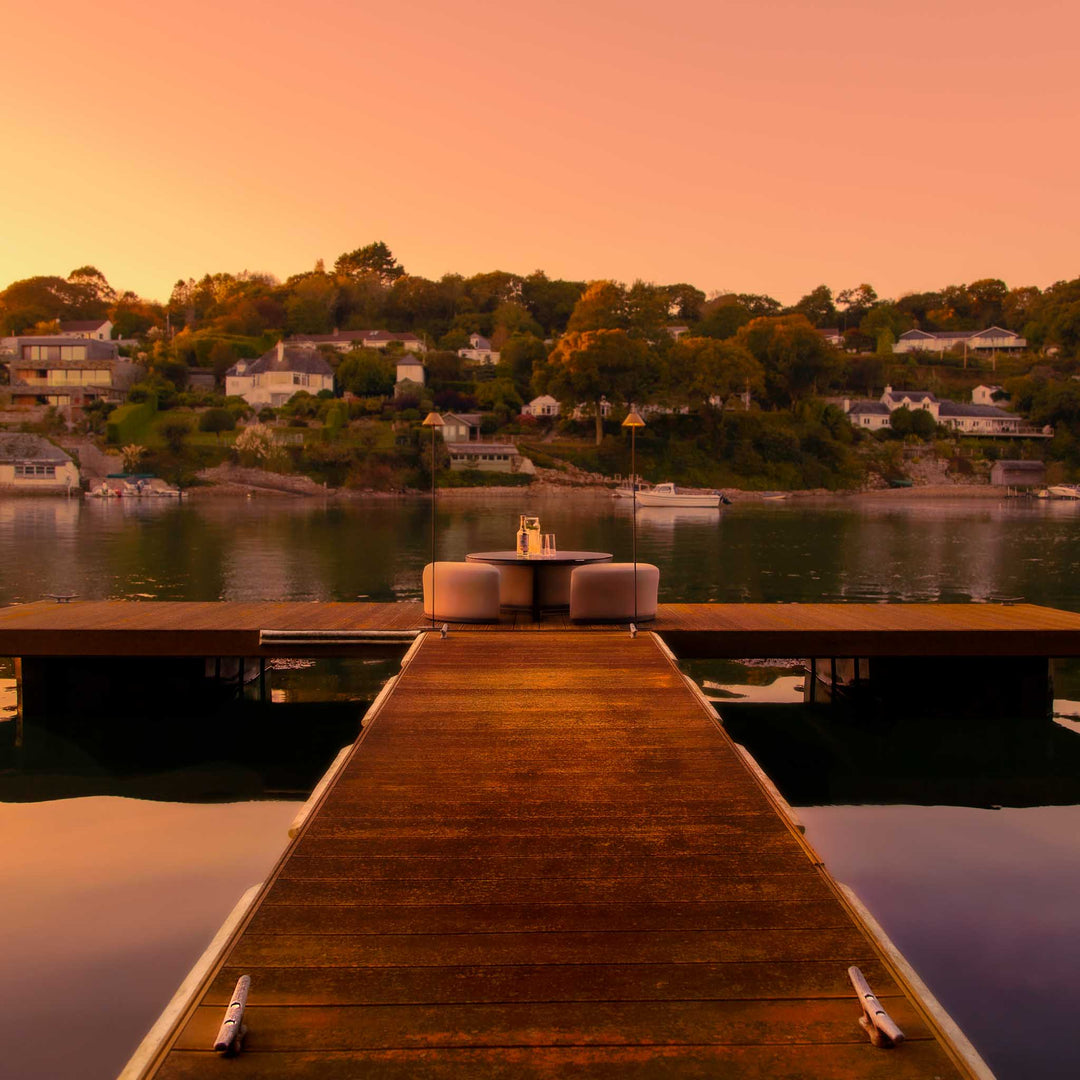 Wooden dock extending into a Cornish estuary  with a sunset over a residential area.