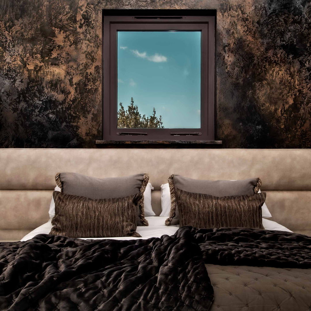 Bedroom with brown bedding and pillows, textured wall, and window view.