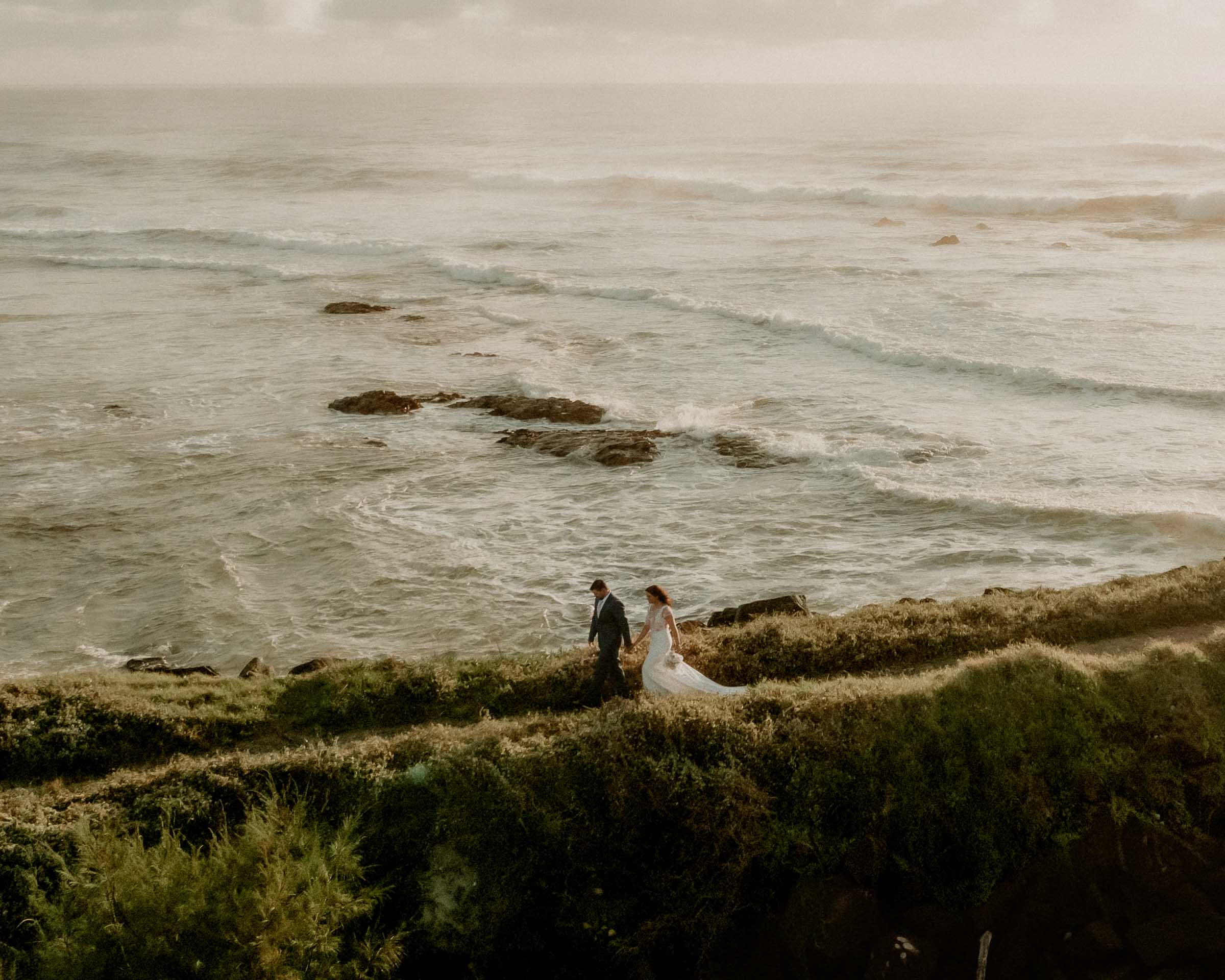 Bride and Groom walking along a coastal path with ocean waves in the background