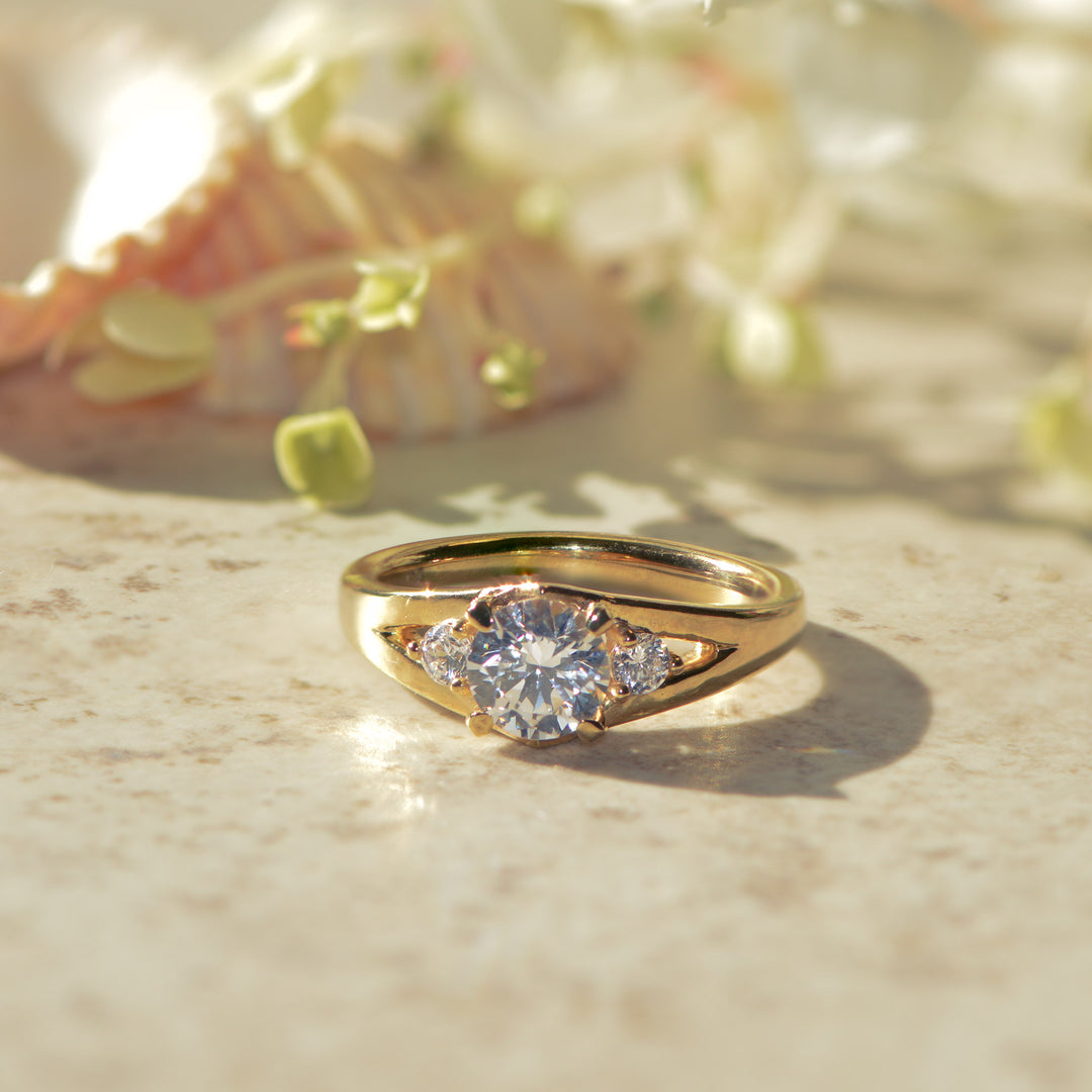 Gold three stone ring with round brilliant cut diamonds on a marble surface with blurred flowers in the background