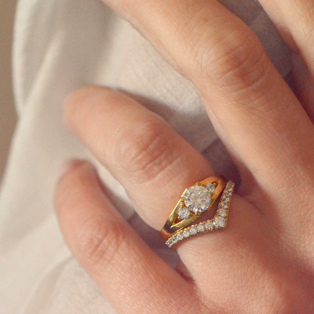 Gold trilogy ring with round brilliant cut diamonds, stacked with a chevron wedding band  on a person's finger against a neutral background