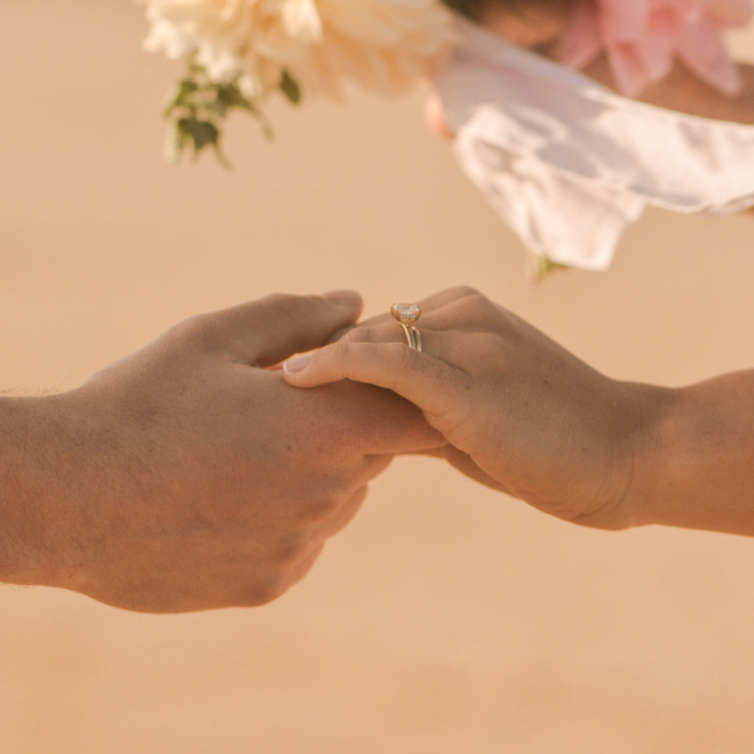 Two hands holding each other, one with a diamond engagement ring, with a blurred background of flowers on a beach.