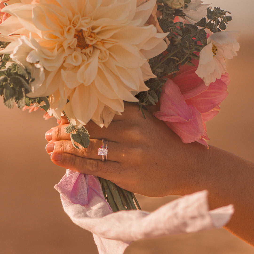 Hand with a radiant cut diamond ring holding a bouquet of flowers with a warm, blurred beach background