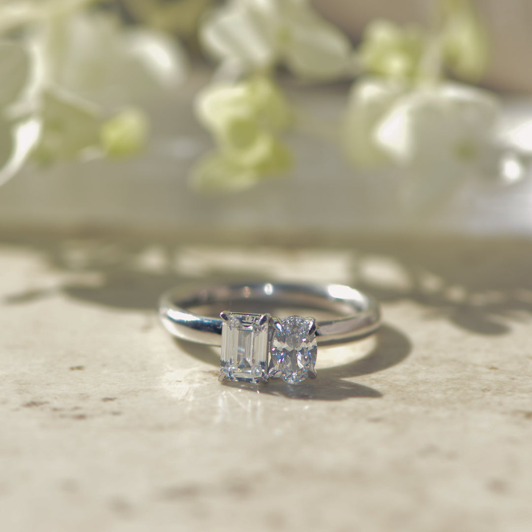 Platinum Toi-et-Moi ring with an emerald and oval cut diamonds on marble with flowers in the background
