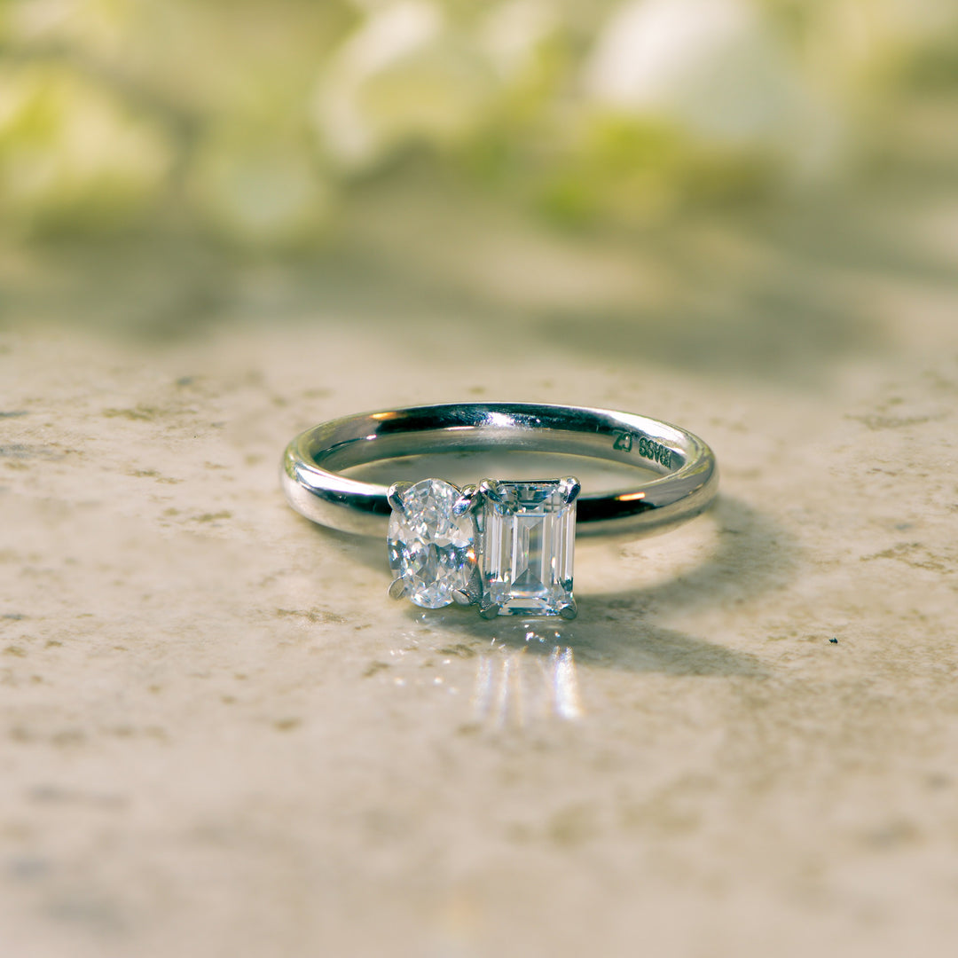 Platinum two stone  ring with an emerald and oval cut diamond on a marble surface