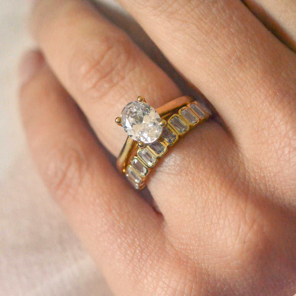 Gold solitaire ring with an oval cut diamond stacked with an art deco wedding band on a hand against a neutral background