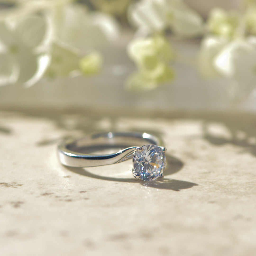 Platinum solitaire ring with a round diamond on a marble surface with flowers in the background