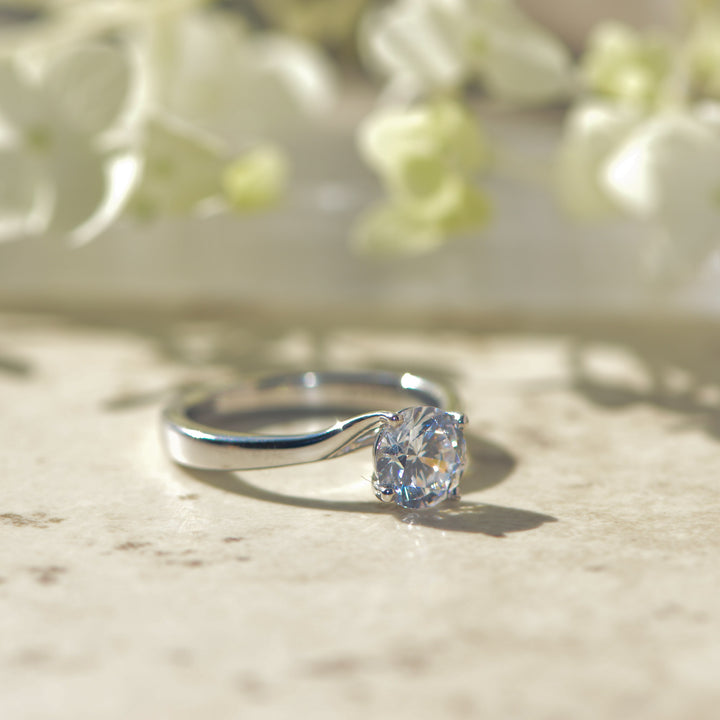Platinum solitaire ring with a round diamond on a marble surface with flowers in the background