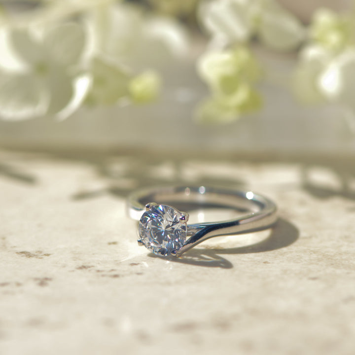 Platinum ring with a round cut diamond on a marble surface with flowers in the background