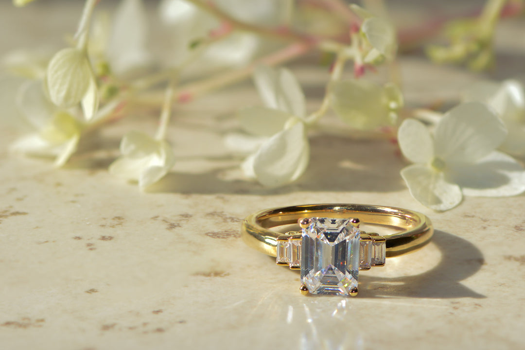 Gold Art Deco emerald ring set with sustainable lab grown diamonds on a light surface with flowers in the background