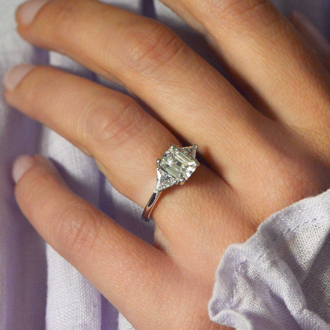 Close-up of a hand wearing an emerald cut diamond trilogy engagement ring with a soft background
